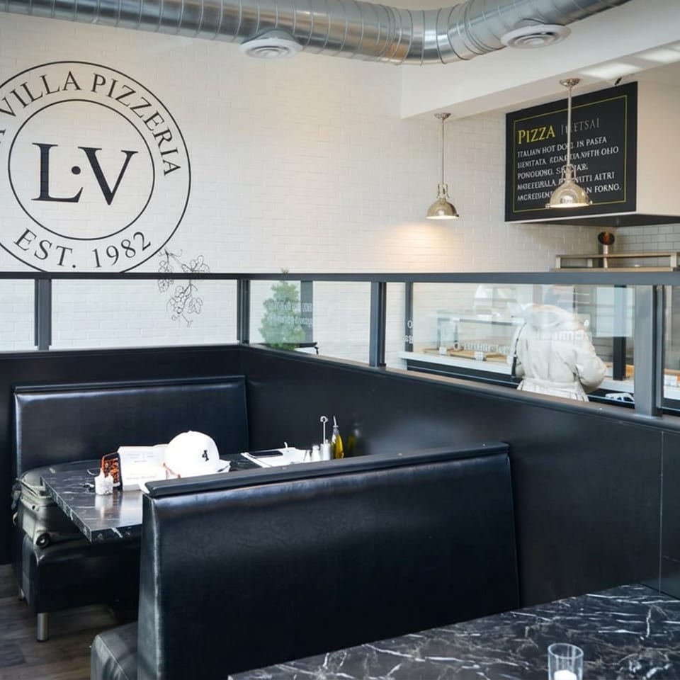 La Villa Pizzeria dining area with black booths, marble tables, exposed ductwork, and an open kitchen counter.