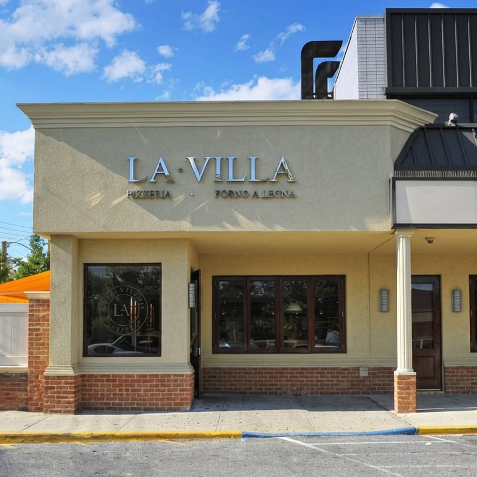 Exterior of La Villa Pizzeria and Restaurant featuring cream walls, brick accents, storefront windows, and a sunny blue sky.