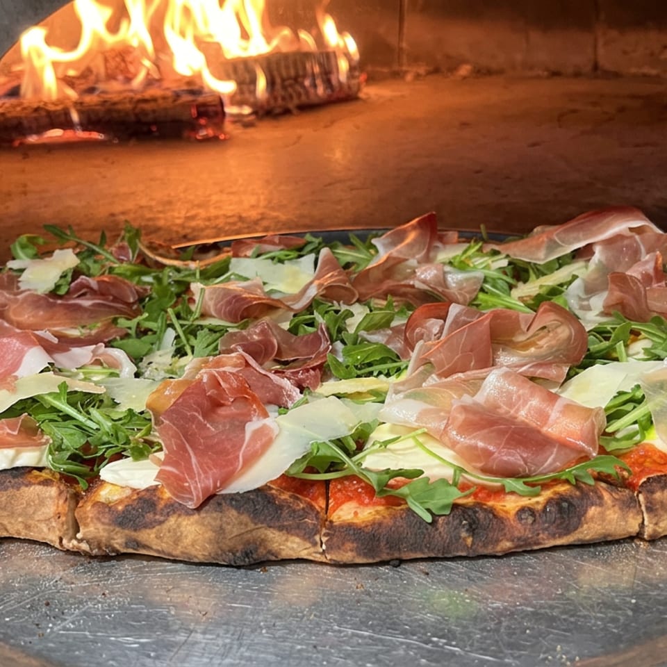 Wood-fired pizza topped with prosciutto, arugula, and shaved cheese on a metal tray beside glowing flames.
