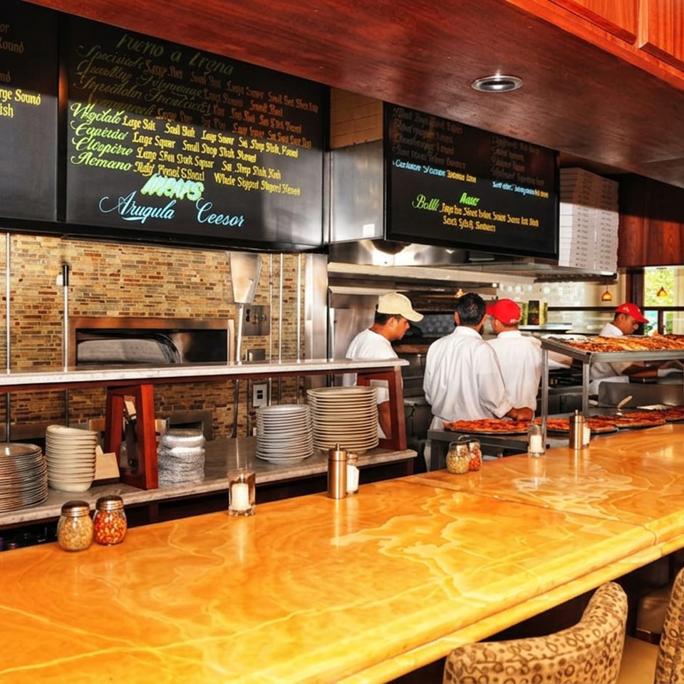 Restaurant kitchen counter with chefs preparing pizzas, stacked plates, menu boards, and warmly lit wooden seating.