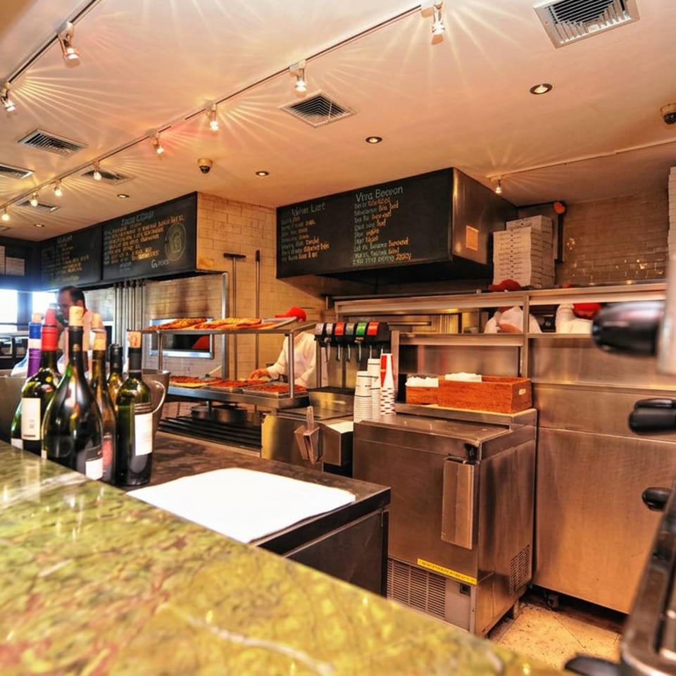 Warmly lit pizzeria counter with wine bottles, pizza ovens, displayed pizzas, kitchen equipment, and chalkboard menus.