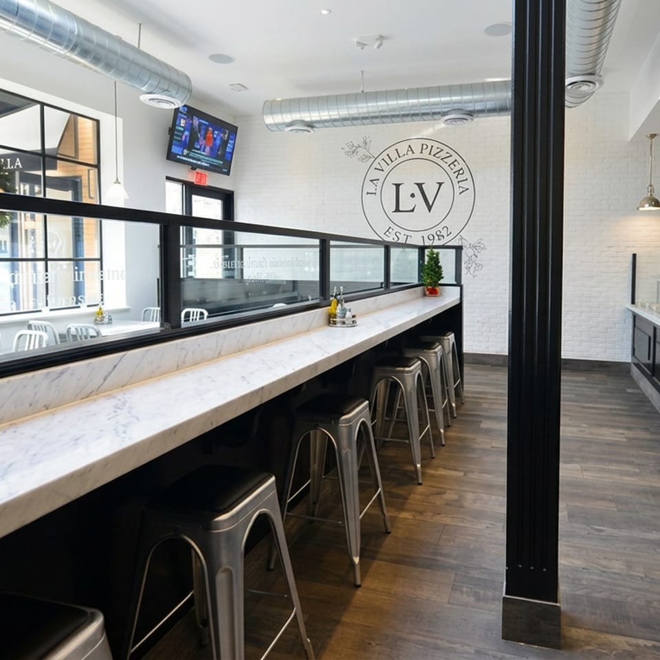 Modern La Villa Pizzeria dining area with marble counters, metal stools, pendant lights, exposed ductwork, and open food counter.