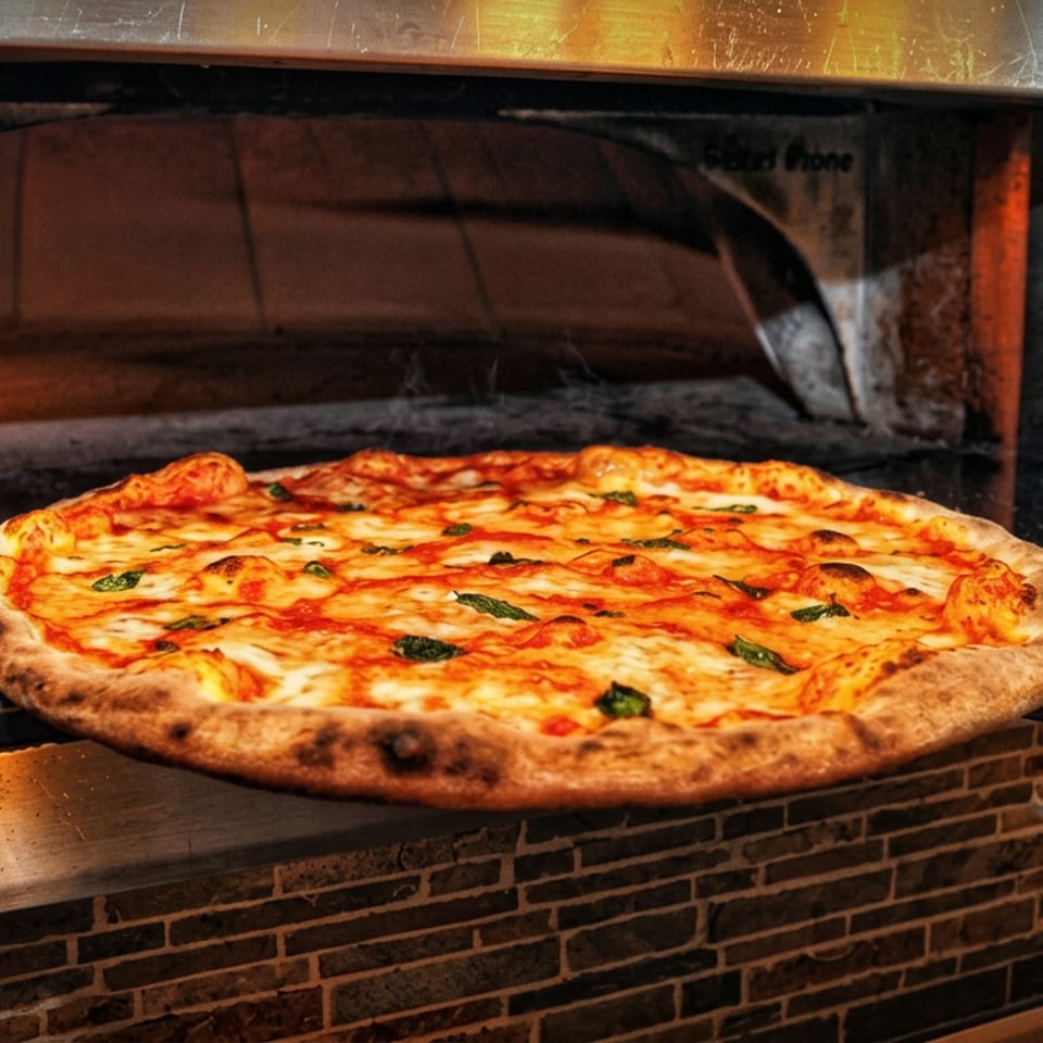 Fresh Margherita pizza with melted cheese and basil rests on a peel in front of a wood-fired oven.