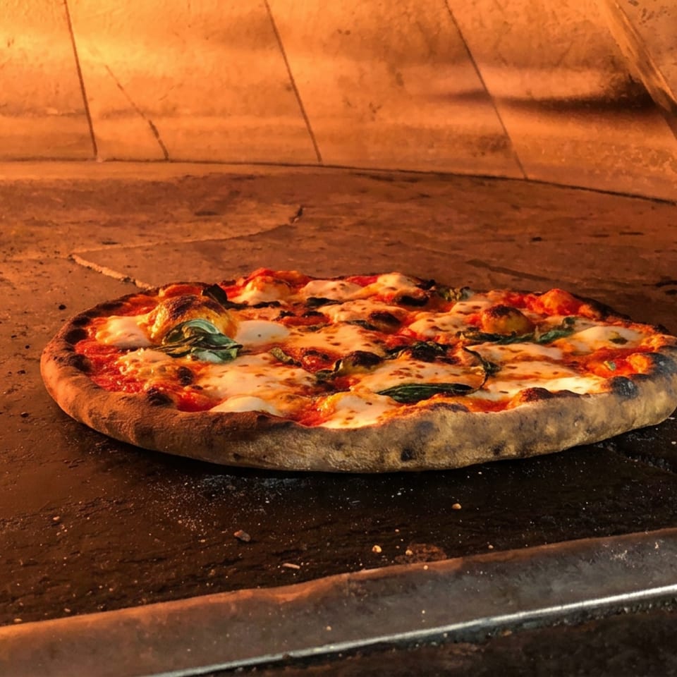 Wood-fired pizza with melted cheese, tomatoes, and charred toppings baking inside a glowing brick oven.