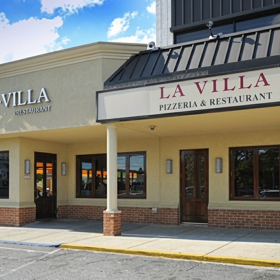 Exterior of La Villa Pizzeria & Restaurant with cream facade, brick accents, glass doors, and prominent signage under a blue sky.