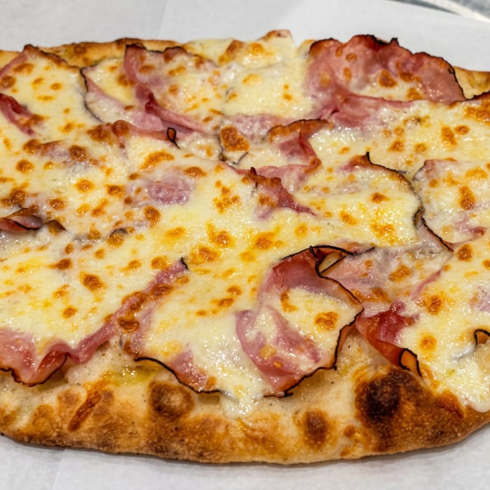Oval, golden-brown flatbread topped with melted cheese and overlapping slices of ham, served on white parchment paper.