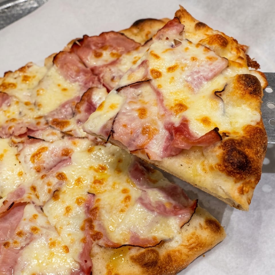 Square pizza topped with ham and melted browned cheese, with a slice lifted by a metal spatula.
