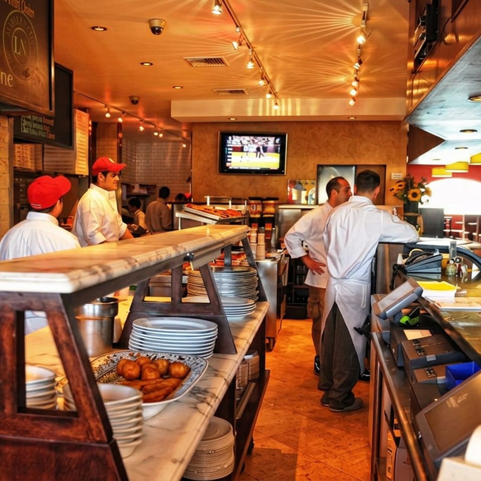 Warmly lit pizzeria interior with chefs working behind the counter, wood-fired oven, stacked plates, and dining tables.